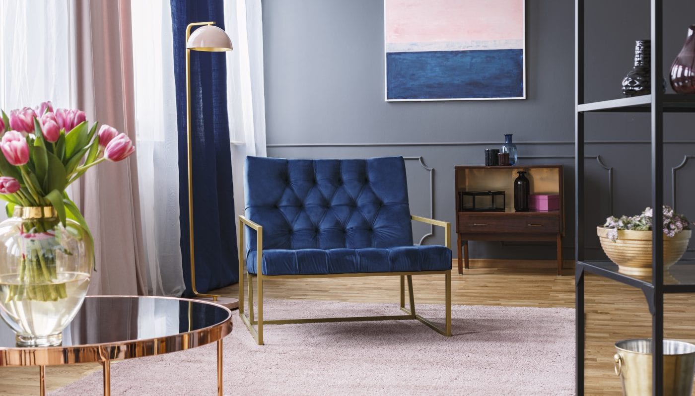 Navy blue armchair next to lamp in sophisticated apartment interior with painting and flowers. Real photo